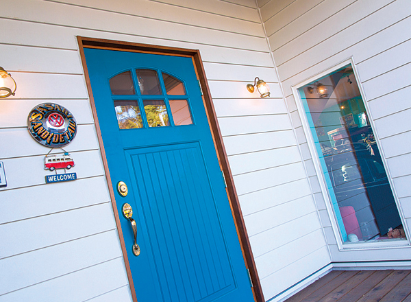 ▲よろい張りのサイディングが施された白い壁面と鮮やかなブルーの玄関ドアにより、アメリカンテイスト溢れる外観に。玄関脇の窓からは、明るく楽しいガレージの様子を窺うことができる